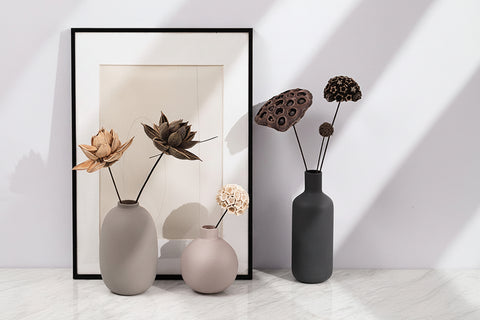 Three vases with dried flowers in front of a framed picture on a white wall.