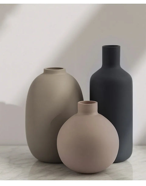 Three matte ceramic vases in neutral tones of taupe, dusty pink, and charcoal gray, arranged on a light marble surface against a soft light gray wall.