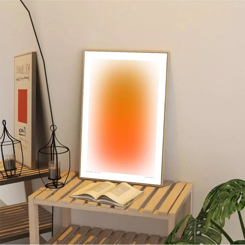 Modern gradient art print in warm orange, on a table beside a book and a plant