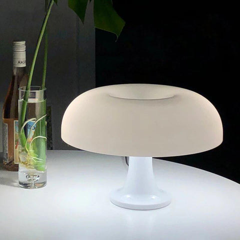 White mushroom table lamp lit on a table beside a glass vase of foliage